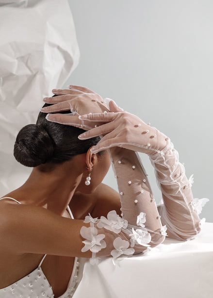Person wearing a white dress with floral gloves and pearl earrings, sitting on a white surface.