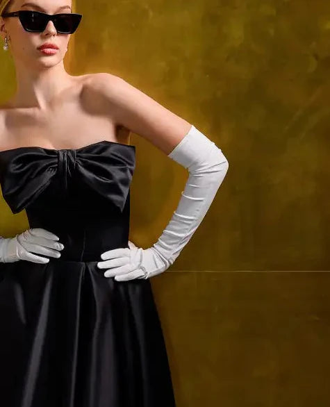 Woman wearing a black dress with a large bow and white gloves against a yellow background