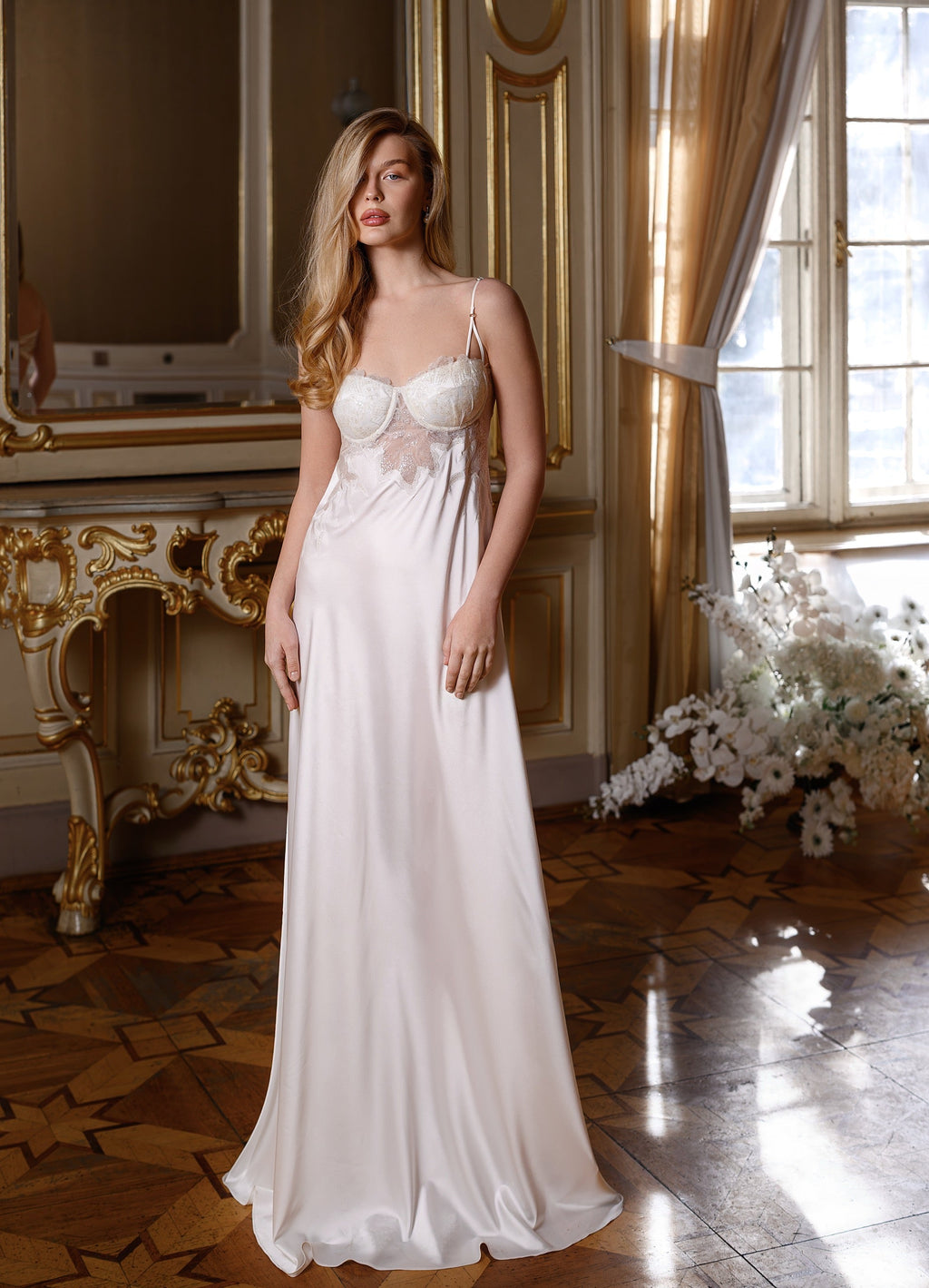 Woman in a white evening gown standing in an elegant room with gold accents and large windows.