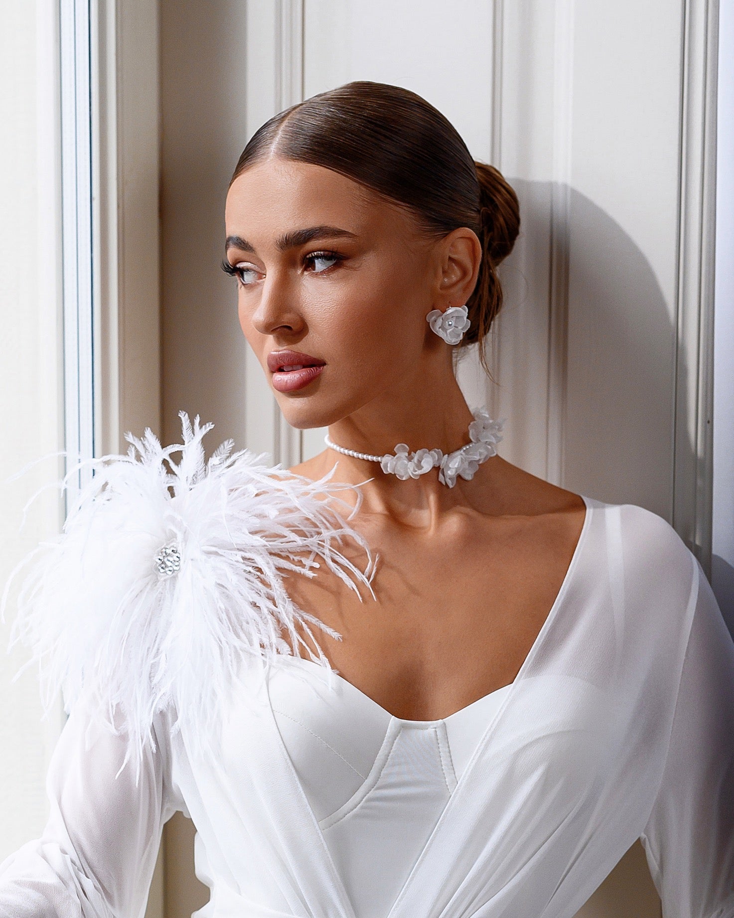 Woman in a white bridal gown with feathered shoulders and jewelry, standing against a neutral wall.